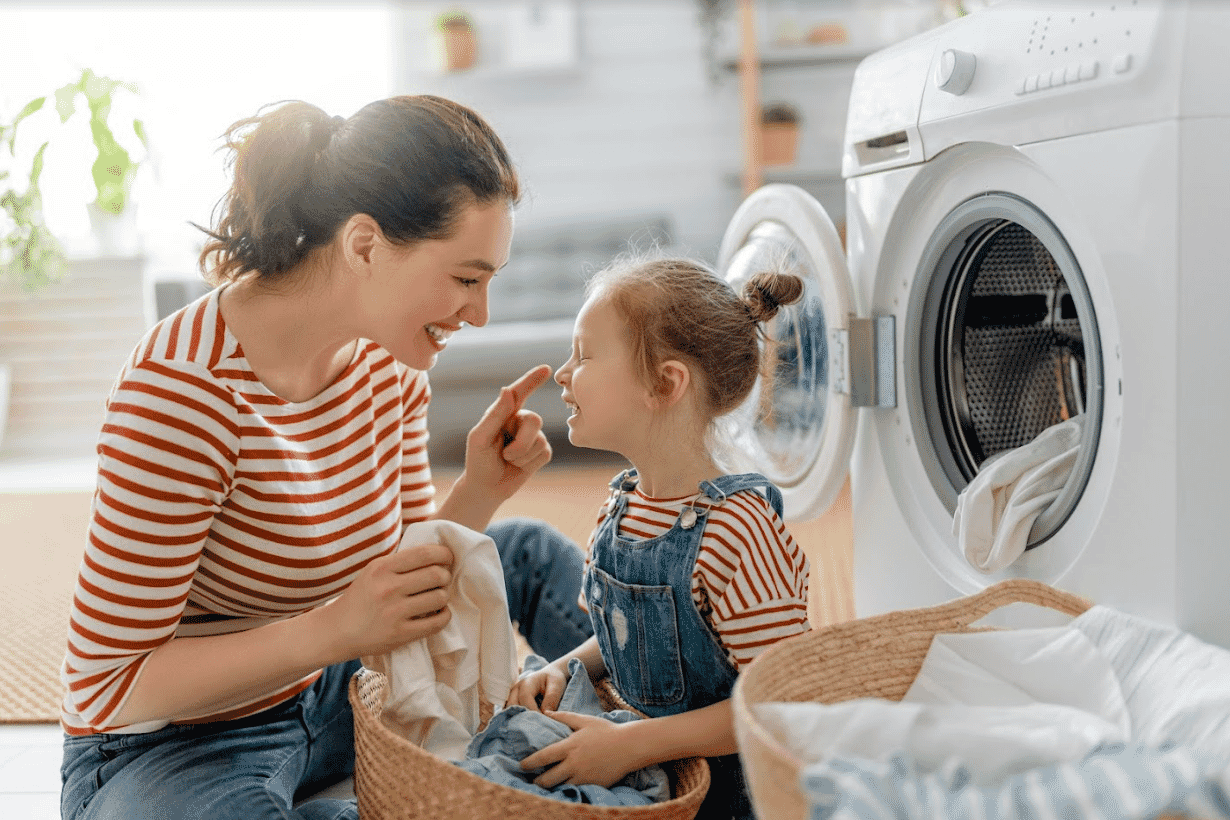 Mother and daughter laugh and smile while sitting on the floor in their laundry with a basket of fresh clothes