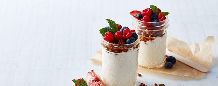 Creamy yoghurt in patterned glass jars topped with granola and fresh raspberries and blueberries on a white surface Image and recipe by Sunbeam