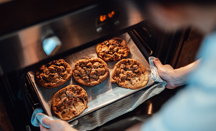 Over the shoulder view of a woman removing a tray of chocolate chip cookies from the oven