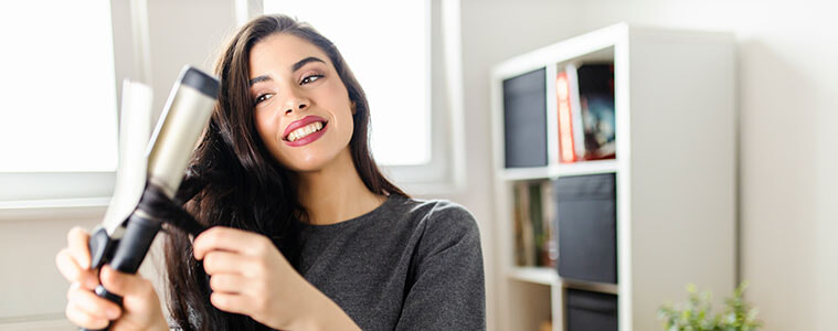 Woman using a curling iron on her long dark hair while sitting at her dressing table