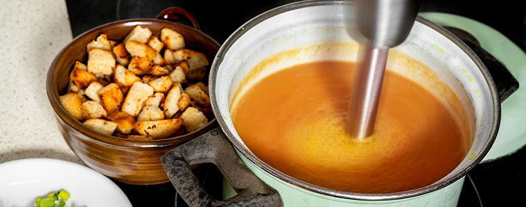 Stick mixer in green pot blending soup next to bowls with chopped vegetables and croutons
