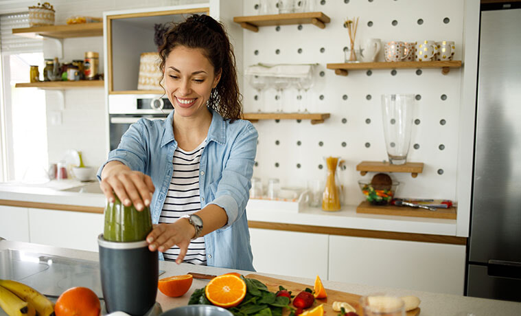 A woman blitzes a green smoothie while standing at her kitchen bench 