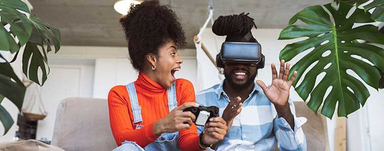 A young man and woman sit on the couch together with the man wearing a VR headset to play a video game