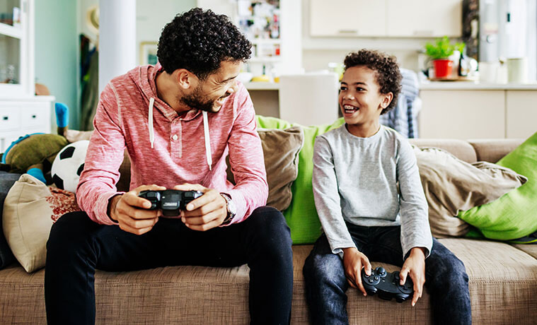 A father and son hold gaming consoles and smile at each other while sitting on the couch during a video gaming session