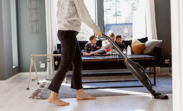 Mother vacuums with a stick vac while her kids play on the couch