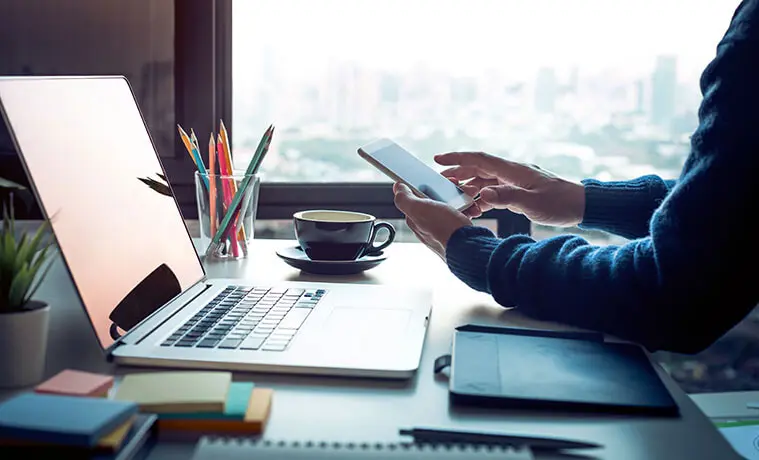 Young man using a smartphone and laptop on his desk at home with coffee cup and notebooks