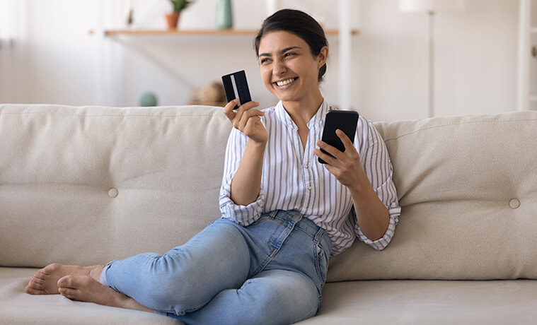 Smiling woman sits on the couch at home with her credit card in one hand and her smartphone in the other