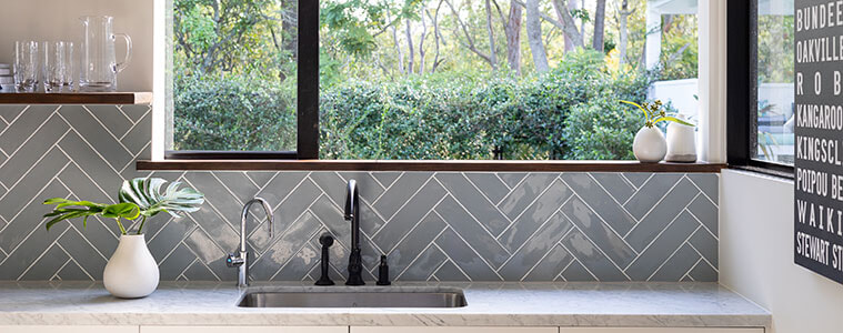 Leafy outlook through a window above a kitchen sink with stylish gooseneck taps marble benchtop and grey splashback tiles in a herringbone pattern 