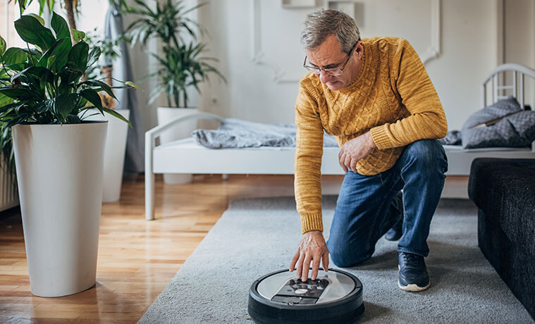 An older man kneels on his living room floor to alter the setting on his robot vacuum cleaner