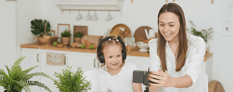 A mother and daughter chat on a video call at the kitchen table with a smartphone positioned in a small tripod