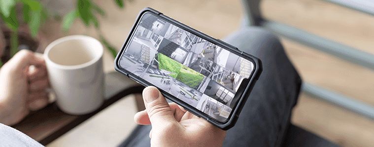 A man sitting in his outdoor area at home with coffee in one hand smartphone in the other viewing footage on its screen from various home security cameras