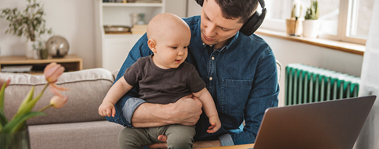 Young father wearing headphones works from home on his laptop with his baby son on his lap