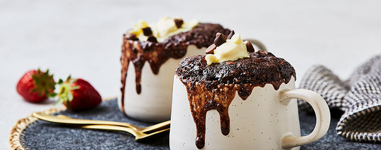 Chocolate cake made with the Bosch Microwave Mug Cake recipe oozing from the top of two white mags on a woven place setting with strawberries and gold teaspoons