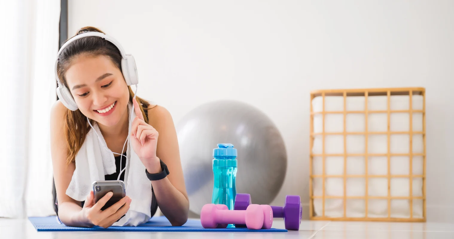 Woman listening to music with headphones and smartphone lying on yoga mat in home gym