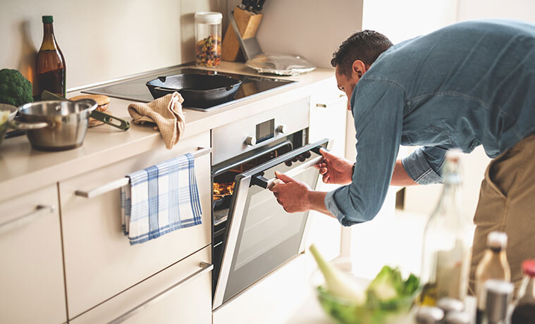 Man in a denim shirt bends to open the under bench oven door to check on dinner