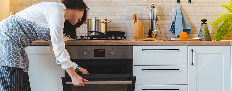 A woman reaches down to open the door of the underbench oven in her modern white kitchen