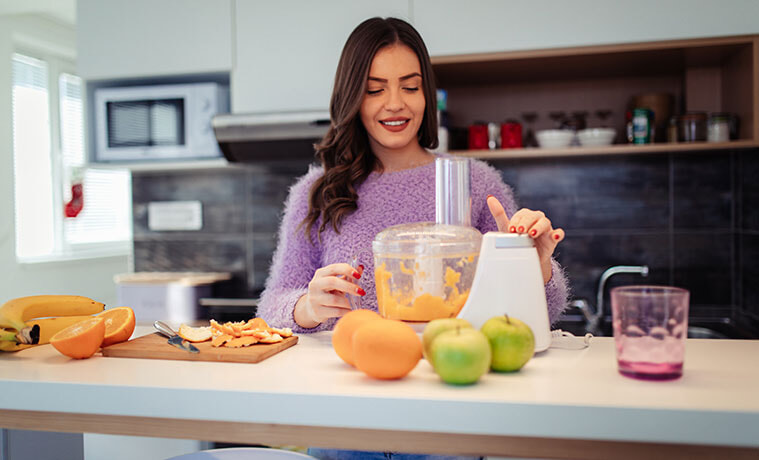 Young woman in a purple jumper juices oranges in a food processor at home