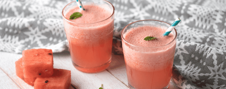 Two glasses filled with watermelon juice with fun straws and chopped pieces of watermelon and mint sprigs at the side