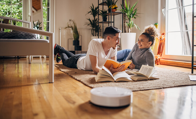 A man and a woman lie on the floor reading books and chatting while their robot vacuum cleans the floor