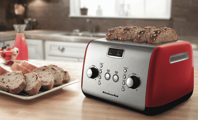 The KitchenAid Artisan 4 Slice Toaster in Empire Red sits on a butcher block kitchen benchtop with 4 slices of thick bread being toasted A plate of toasted bread sits to the left of the toaster