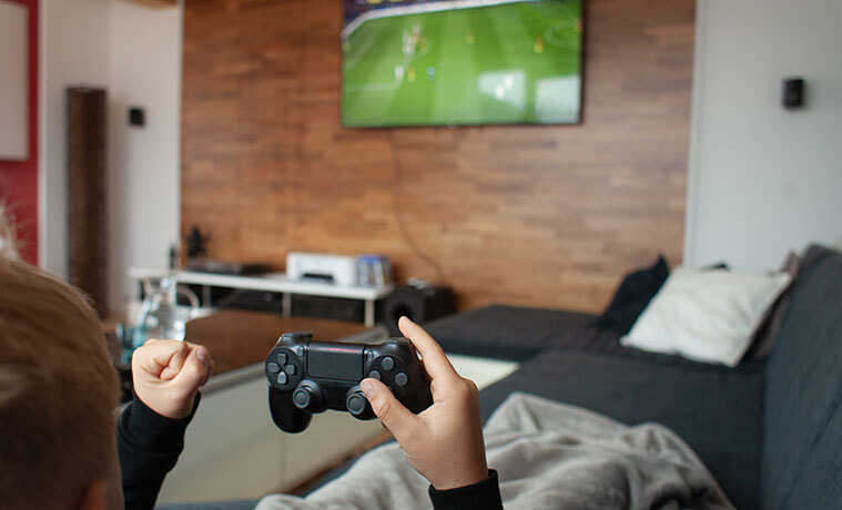Overtheshoulder view of a boys hands holding a gaming console in front of a TV screening a football game