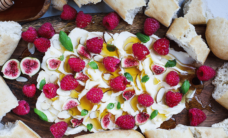 Creamy butter on a wooden board topped with honey and herbs with a honey dipper and slices of bread