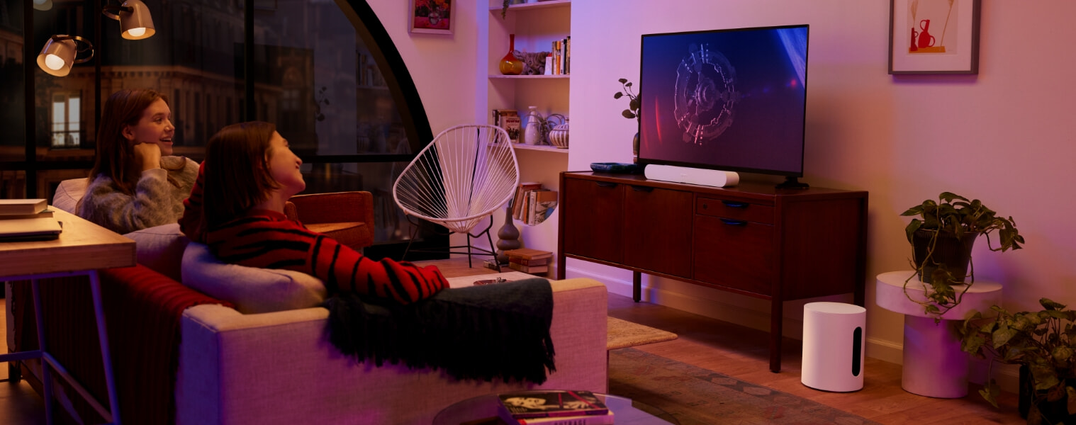 Two woman sitting on their couch watching a movie that is playing through their Sonos Sub Mini speaker 