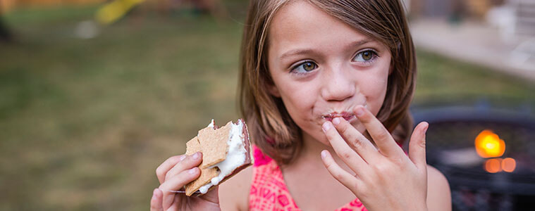 Girl looking away while eating a smore in her yard with a barbecue grill in the background