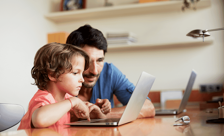 A father and his young son sit together in front of a laptop on a desk in their home office