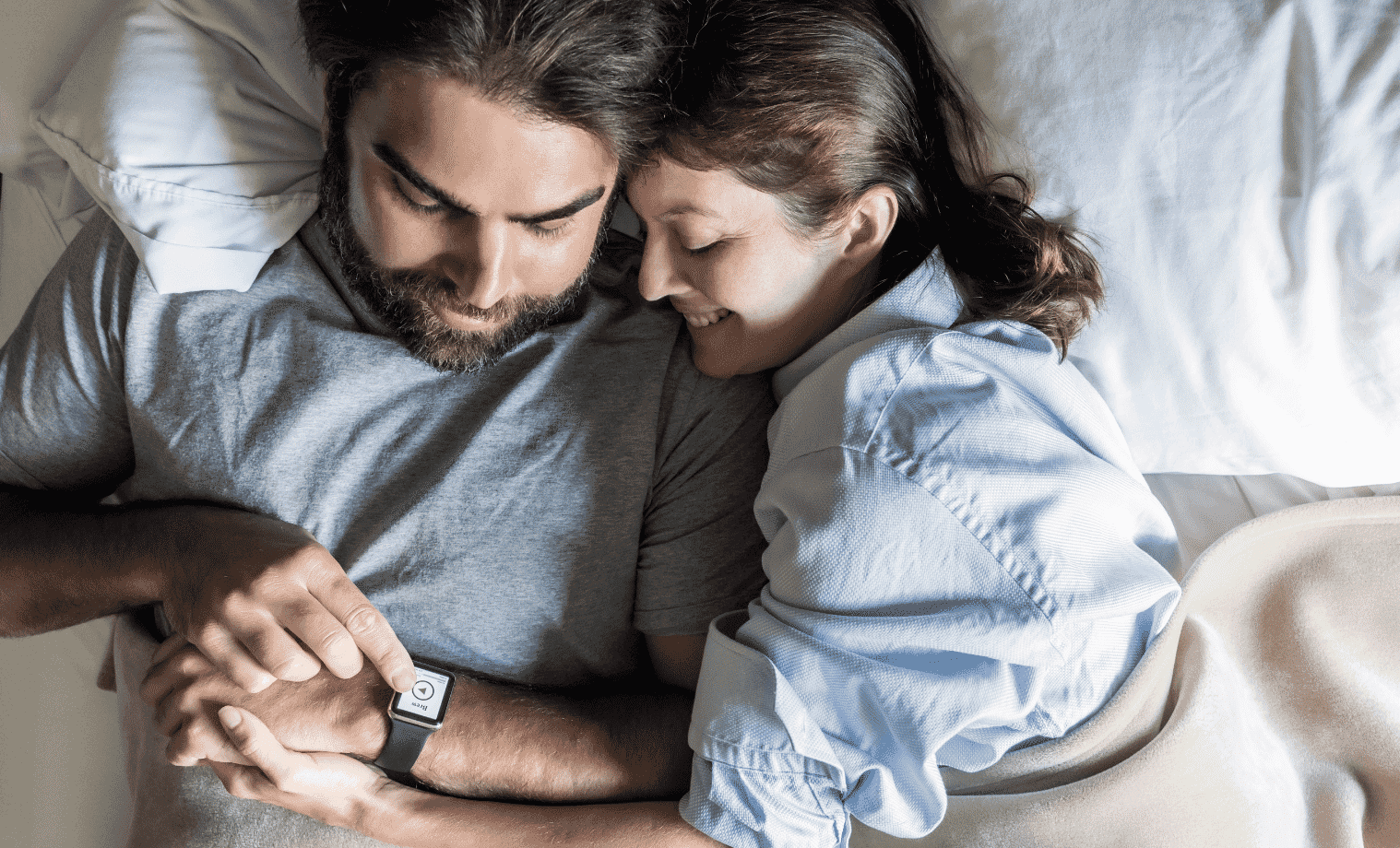 A man and woman lay in bed together and examine the mans smart watch