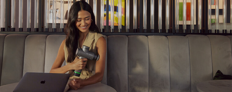 A woman sits at a table with her laptop and uses and Hyperice Hypervolt massage gun on her arm