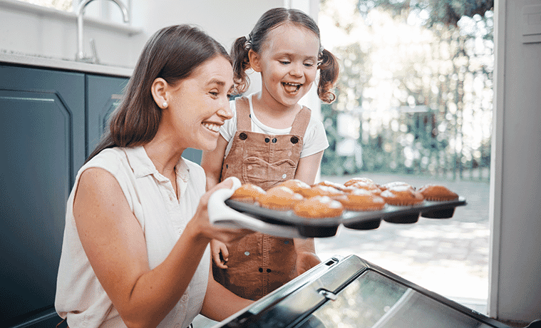 A mother and daughter pull a tray of muffins from the oven
