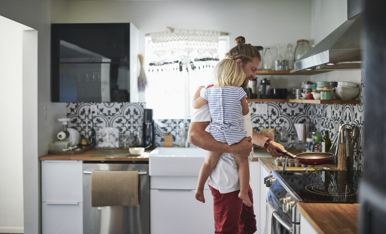 A father carrying his toddler daughter places an empty frying pan on an induction cooktop in his kitchen