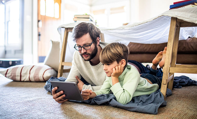A father and son watch a TV show on their digital tablet while sitting under a homemade fort in their living room