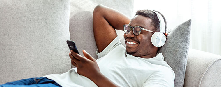 A young man reclines on a couch with headphones on and smartphone in hand watching a game on the screen