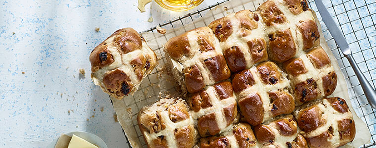 Hot cross buns on a tray lined with baking paper with mini white Easter eggs on top sitting on a timber table with a cup of tea