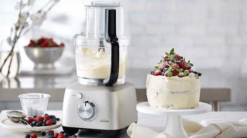 Sunbeam food processor with cream in it placed next to a delicious cake with strawberries on top