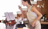 A young woman leans over a pot on her kitchen cooktop and prepares to sample the sauce she is making