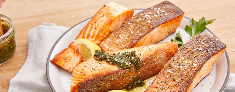 Salmon pieces on a plate sitting on a white napkin on a wooden table a hand spooning a marinade onto the fish with a spoon