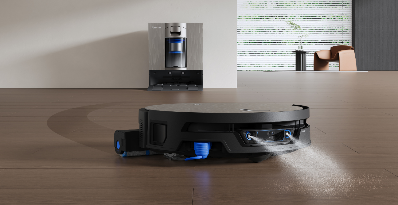 Close-up of a black ECOVACS robot vacuum cleaning a wooden floor, spraying a fine water mist from its underside with its extendable side mop arm reaching out to the left. Its tall silver docking station sits against the wall behind it in a bright, modern living room.