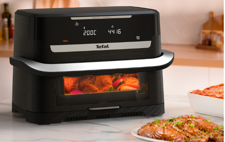 Tefal Infra Heat air fryer with a viewing window showing food cooking inside, next to a plate of glazed salmon and a baked dish on a marble bench.