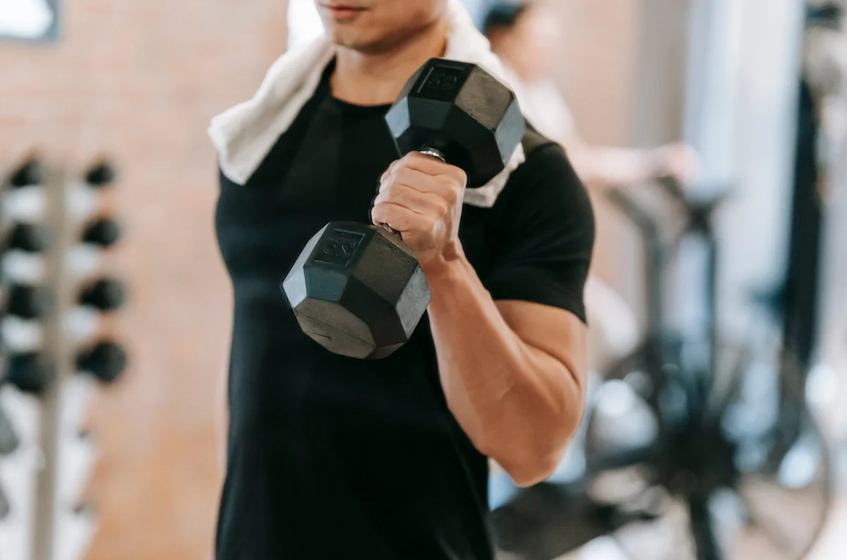 Man in a black t-shirt with a towel over his shoulder curling a dumbbell in a gym, weight rack blurred behind him