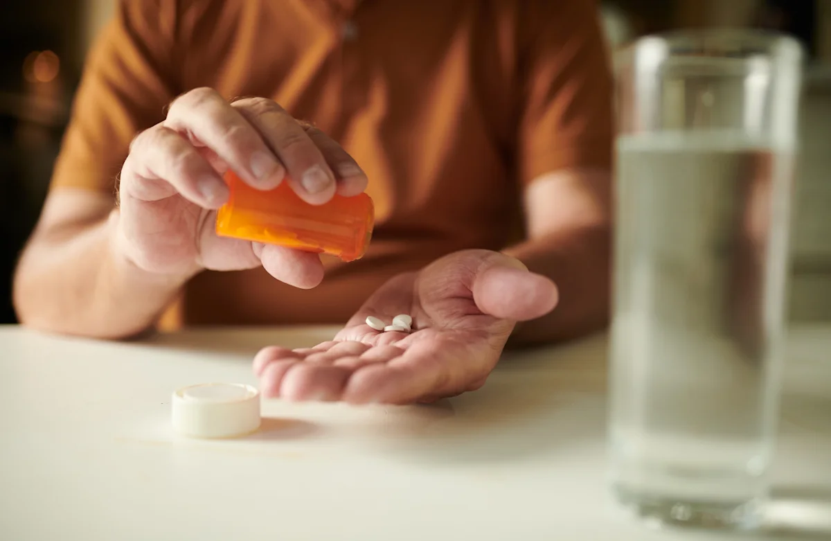 Man tipping two white tablets from an amber pill bottle into his palm, with a glass of water on the table