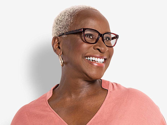 A lady with short cropped grey hair wearing a pink top and dark frames glasses