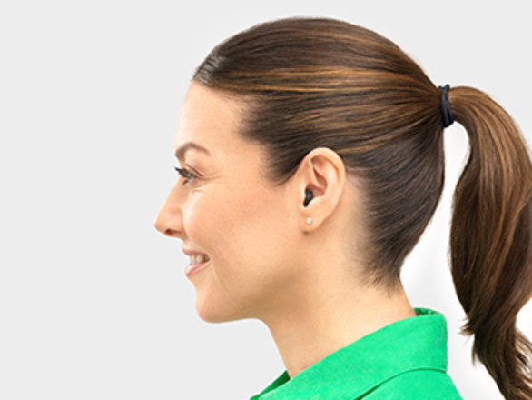 A woman in a green shirt with long brown hair in profile and wearing a hearing aid