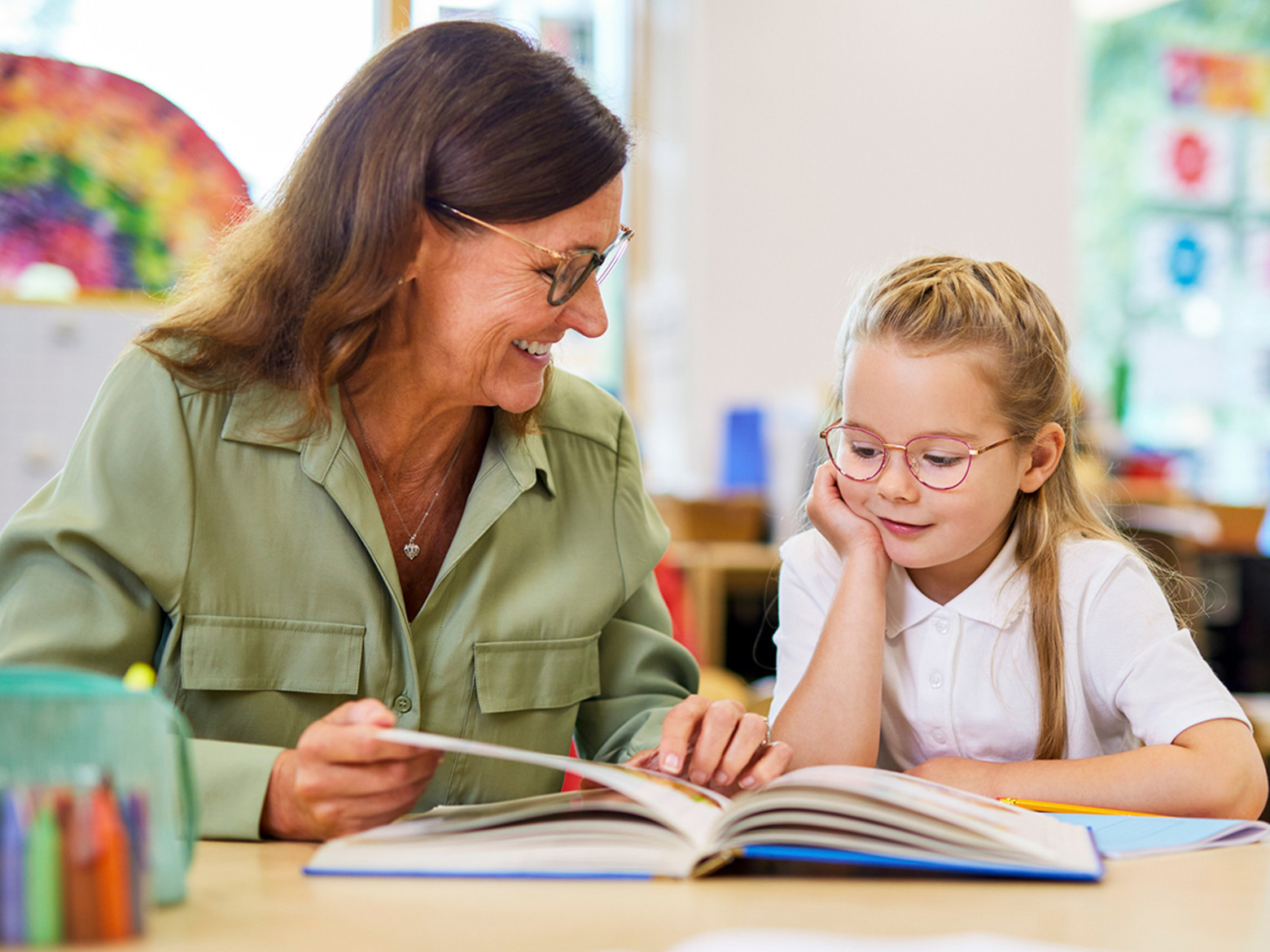 A female teacher reads a book with a young girl who is wearing glasses
