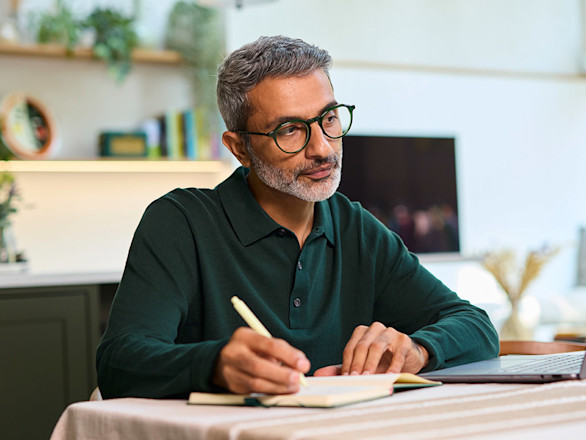 Man in dark green shirt and wearing glasses working at a table with his laptop while making notes.