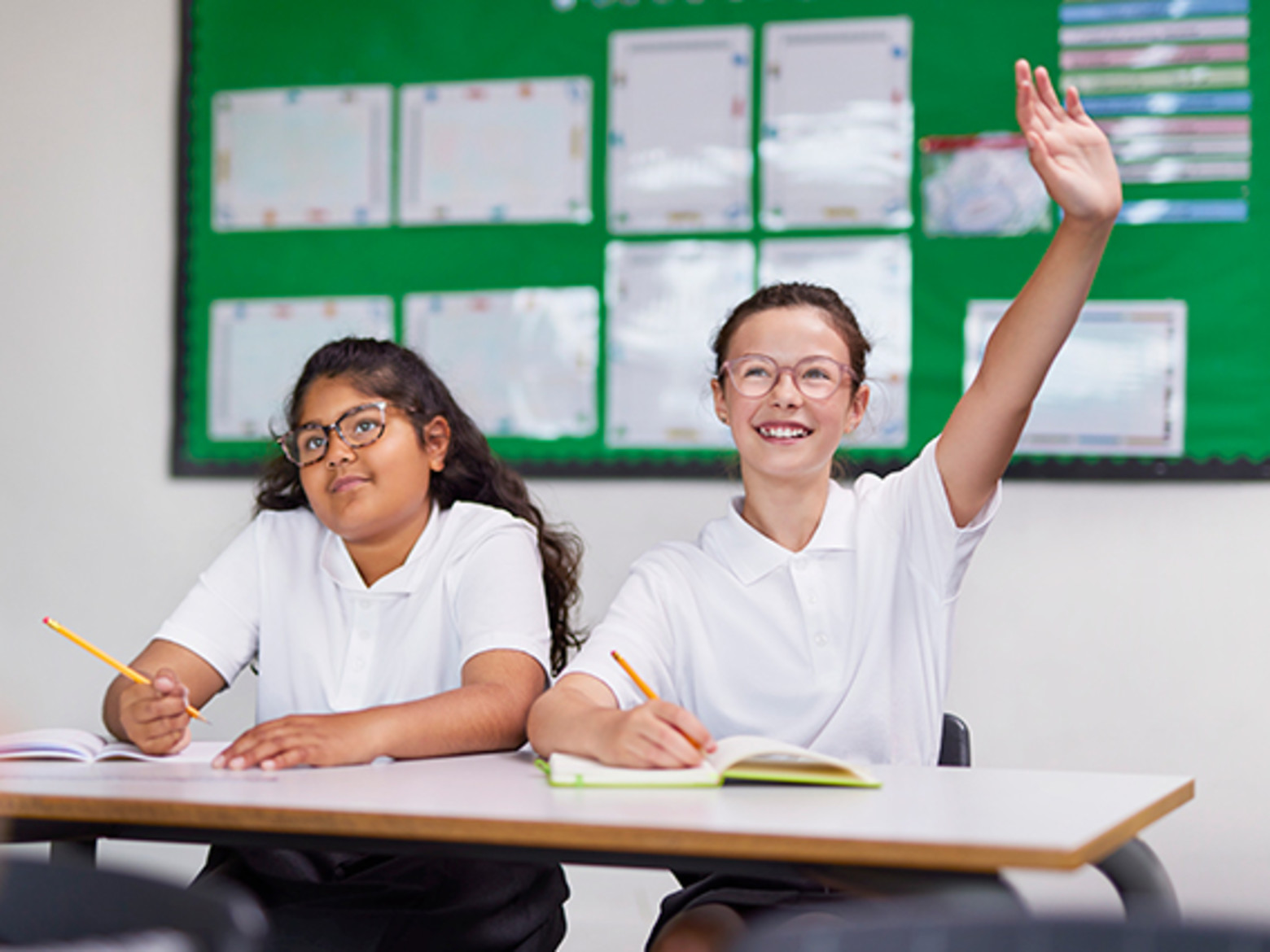 Two girls in a class room, one is writing in an exercise book, the other has her hand raised