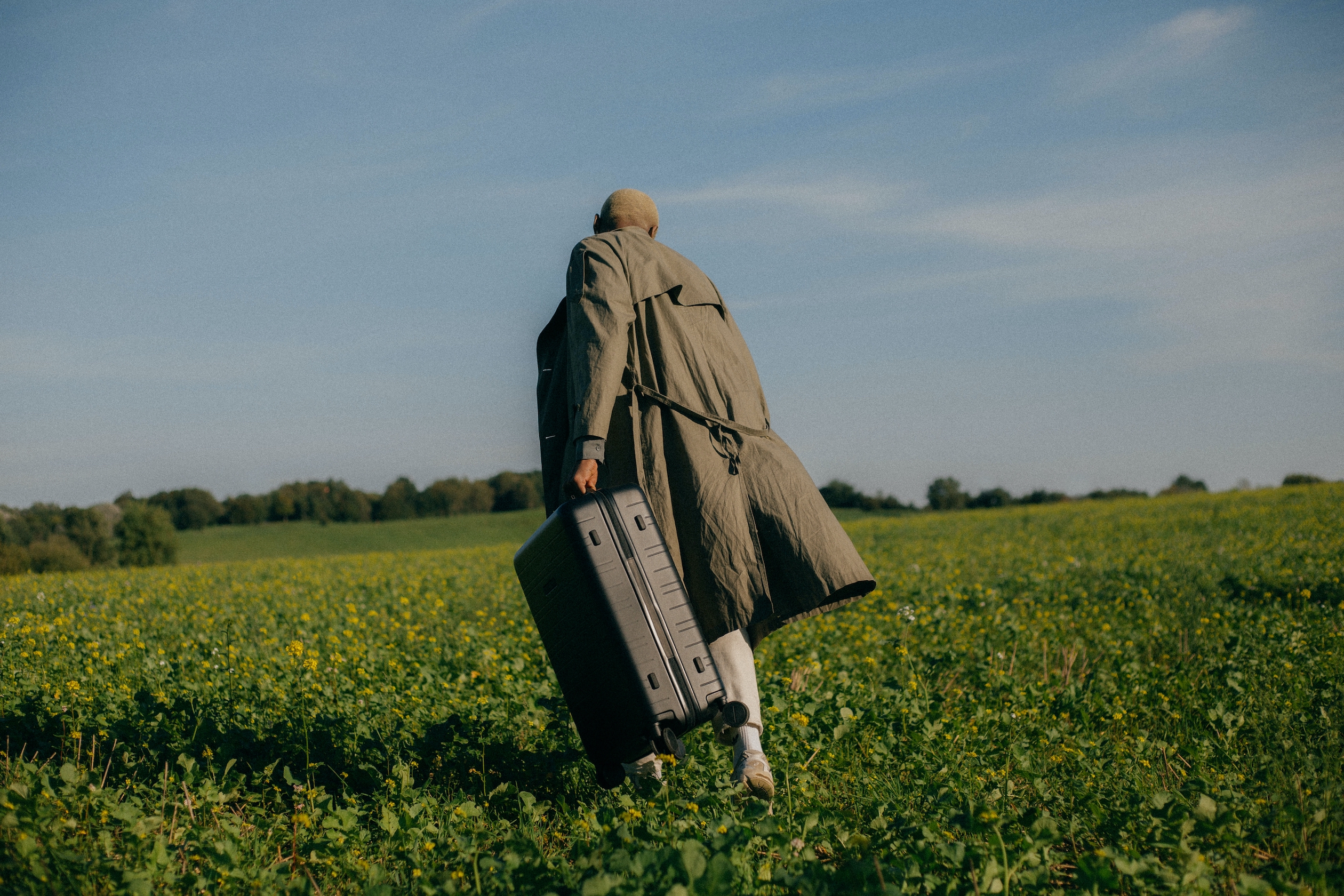 Photo of a person walking through a lush open field wearing a trench coat and carrying a suitcase.