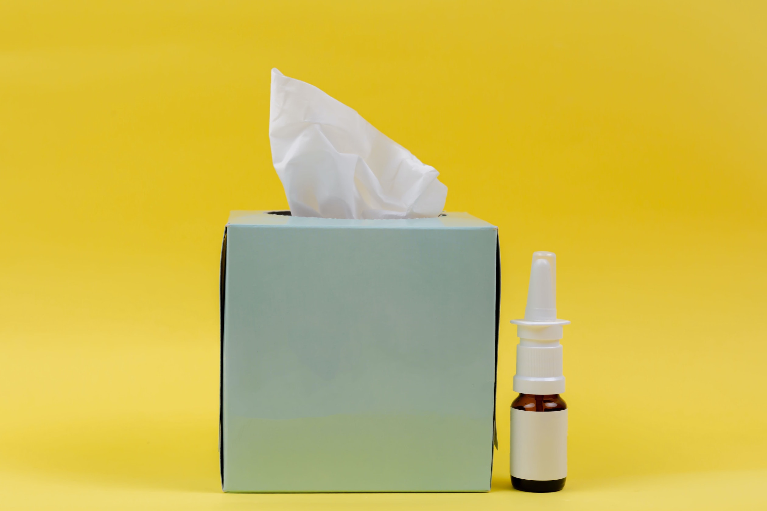 Photo of a box of tissues and a bottle of nasal spray in front of a bright background.